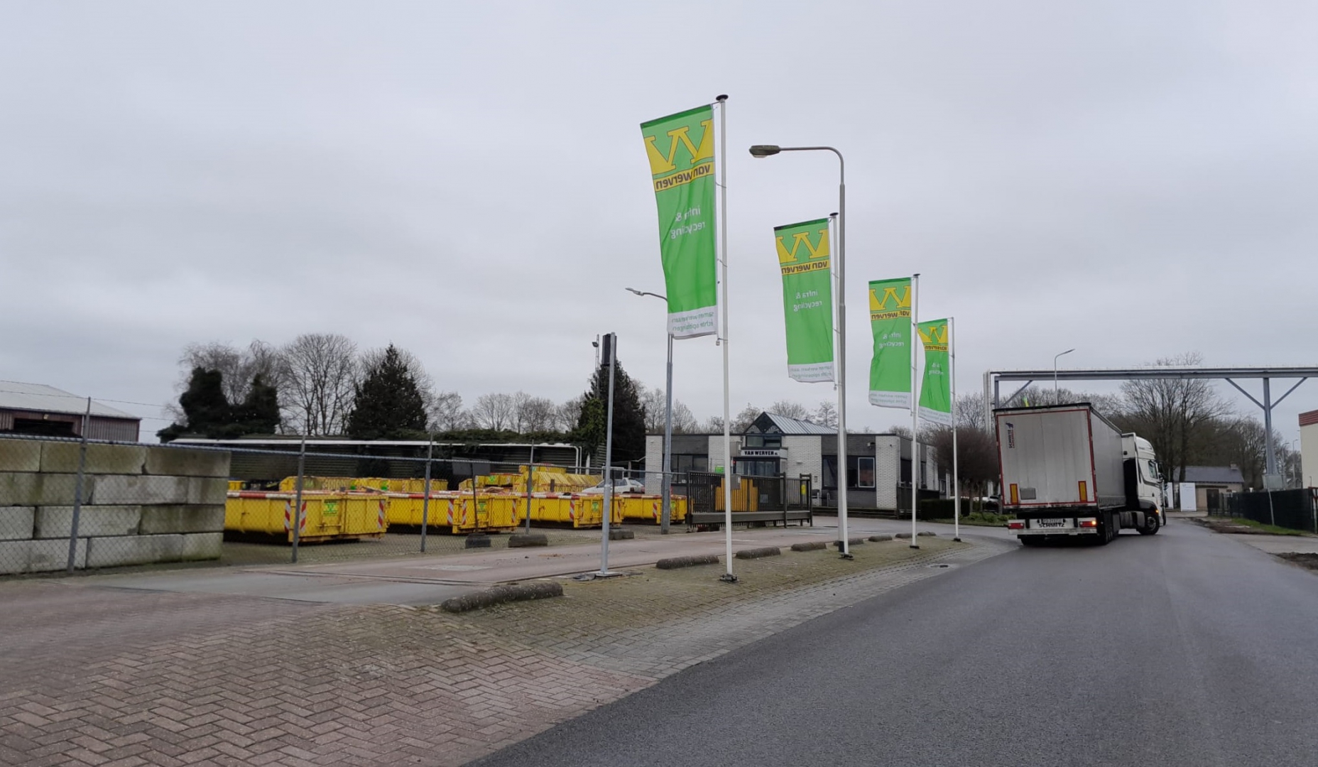 Vier Van Werven-vlaggen langs de weg bij de vestiging Balkbrug, met gele containers achter het hek en een passerende vrachtwagen