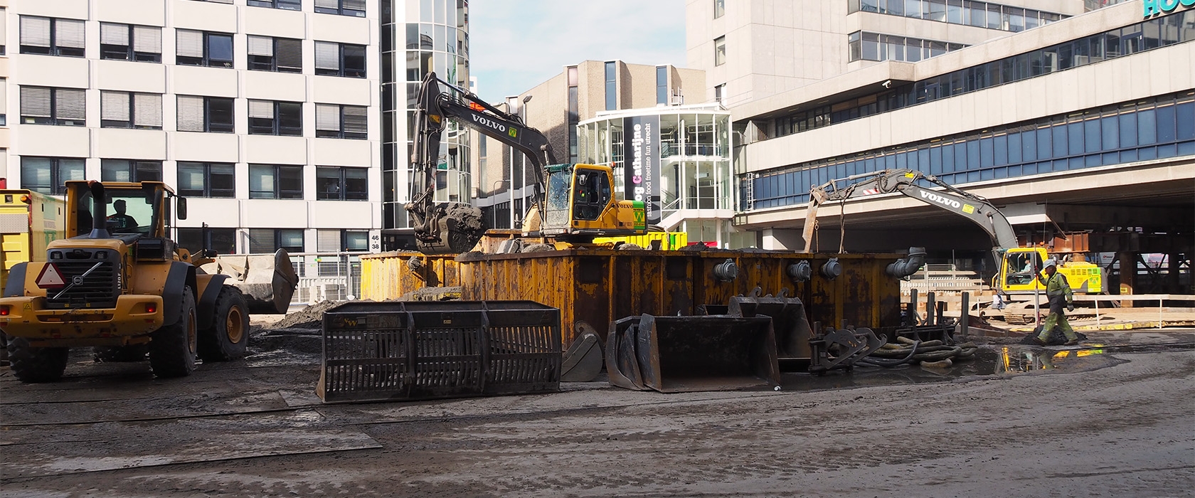 Bouwterrein voor de ingang van Hoog Catharijne: een wiellader, een midigraver op de gele hulpconstructie en een rupskraan met lange giek van Van Werven