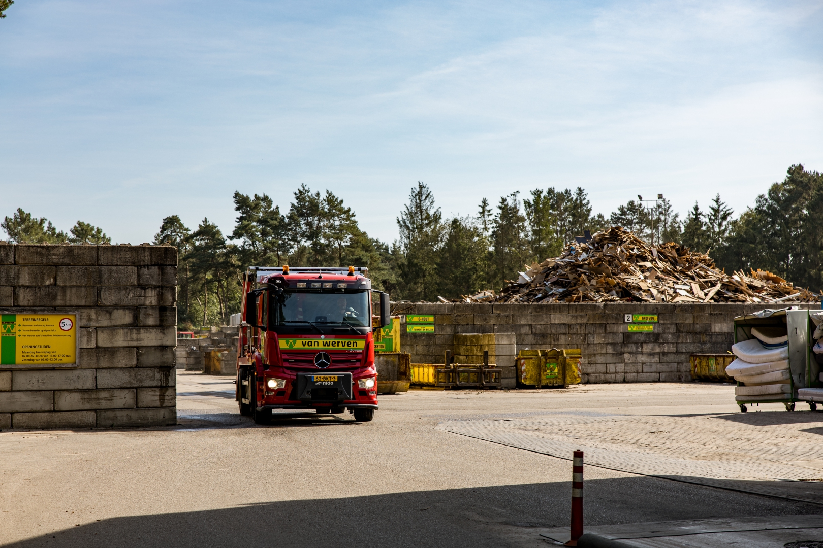 Een rode Van Werven-vrachtwagen rijdt de poort van de recyclinglocatie uit, met een berg houtafval op de achtergrond