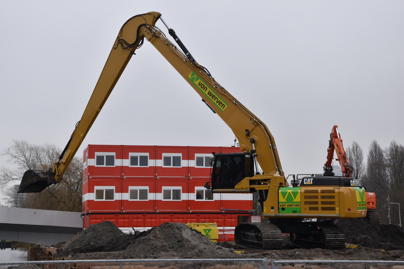 De rupskraan ECO Longreach graaft met de lange giek naast een bouwkeet van stapelcontainers
