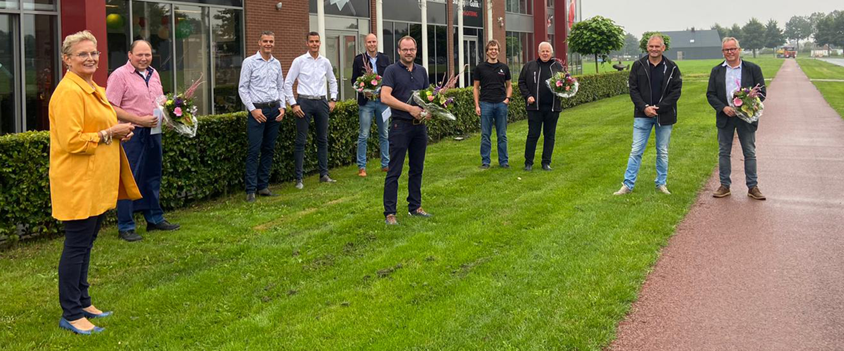 Tien mensen met bloemboeketten staan op afstand van elkaar op het gras voor het kantoorpand