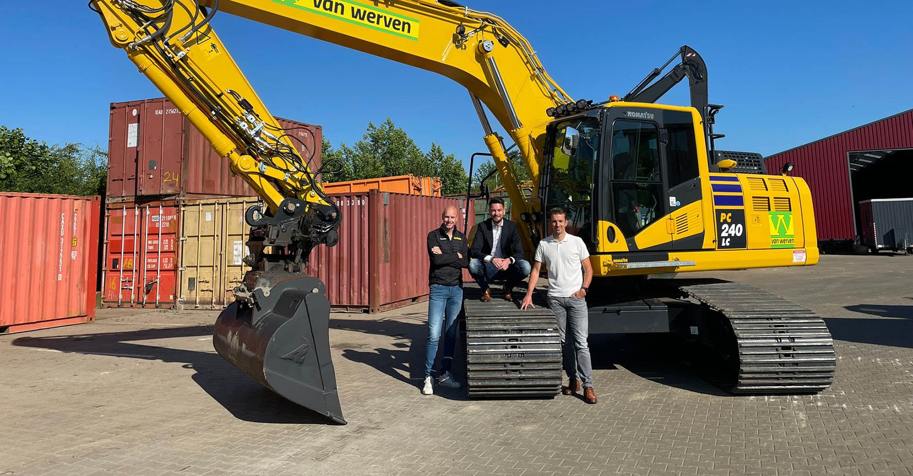 Drie mannen poseren op de rupsband van een gele Komatsu PC240-graafmachine van Van Werven tussen zeecontainers