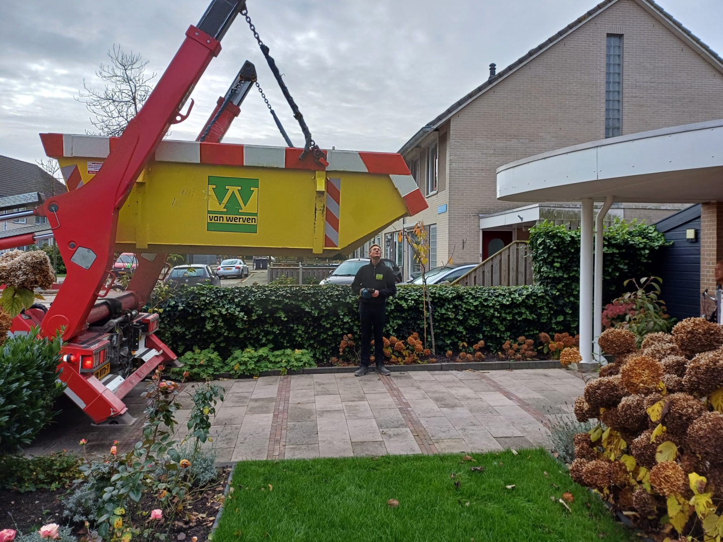Een medewerker met afstandsbediening plaatst met de kraanwagen een gele Van Werven-container in een voortuin