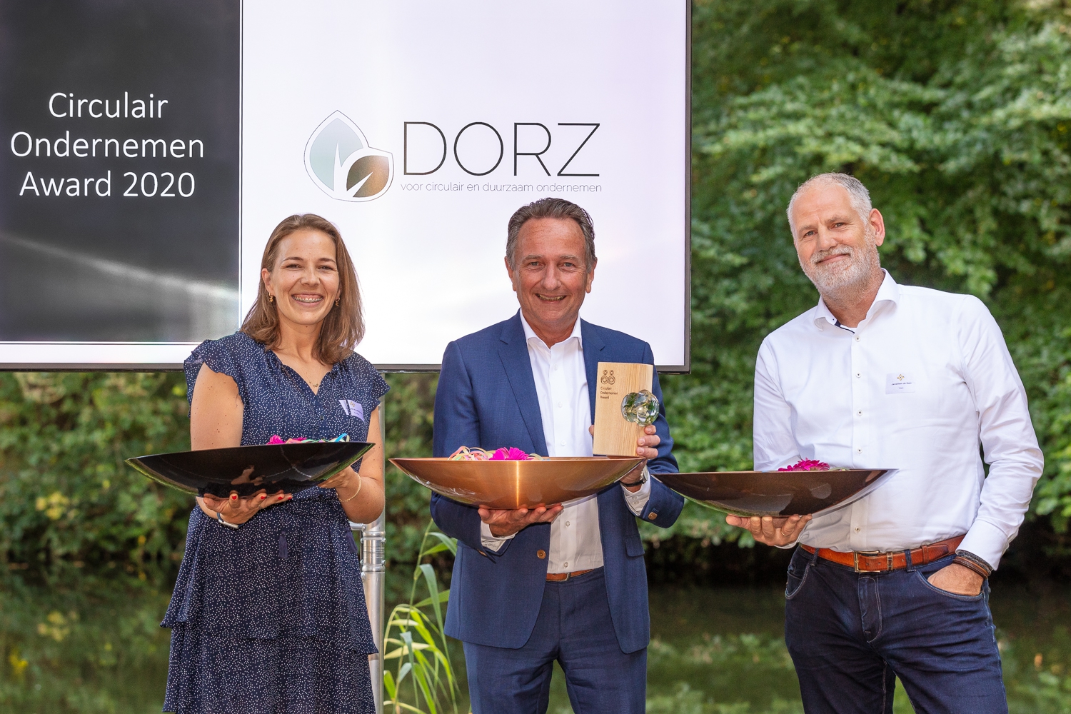 Drie prijswinnaars met schalen en de houten Circulair Ondernemen Award, voor het scherm van organisator DORZ