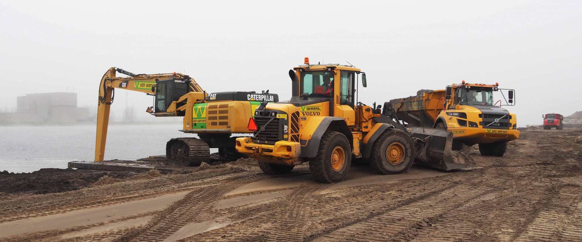 De Cat met lange giek, een Volvo-wiellader met rijplaat en een dumper achter elkaar op de zandbaan langs het water