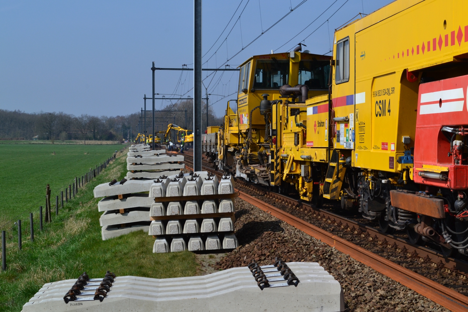 Stapels betonnen dwarsliggers langs het spoor, met een gele spoorwerkmachine ernaast en weiland op de achtergrond