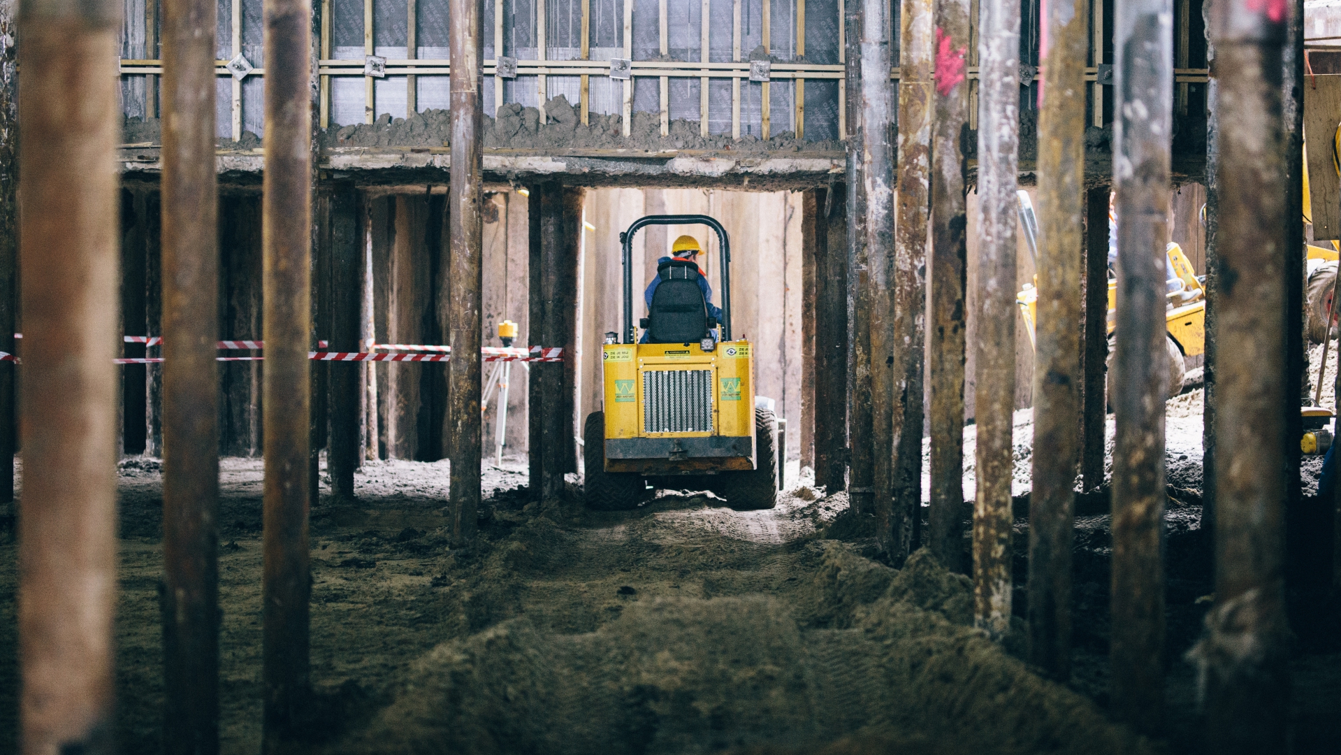 Een klein kiepwagentje rijdt met een lading grond tussen de rijen stalen buispalen onder het gebouw door